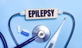 On a light gray background, a stethoscope, an electronic thermometer, pills, a wooden block with the text EPILEPSY. Medical