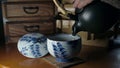 Light Japanese tea is poured with a wrinkled hand from a dark brown clay pot into an old chipped tepid white porcelain cup