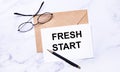 On a light marble background, a craft envelope, glasses, a pen and a white card with the text FRESH START