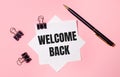 On a light pink background, black paper clips, black pen and white note paper with the words WELCOME BACK. Flat lay Royalty Free Stock Photo