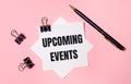 On a light pink background, black paper clips, black pen and white note paper with the words UPCOMING EVENTS. Flat lay Royalty Free Stock Photo