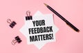 On a light pink background, black paper clips, black pen and white note paper with the words YOUR FEEDBACK MATTERS. Flat lay Royalty Free Stock Photo