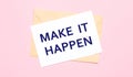 On a light pink background - a craft envelope. It has a white sheet of paper that says MAKE IT HAPPEN