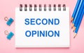On a light pink background, light blue pencils, paper clips and a white notebook with the text SECOND OPINION Royalty Free Stock Photo