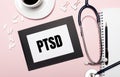 On a light pink background, a notebook with a pen, stethoscope, white pills, paper clips and a sheet of paper with the text PTSD.