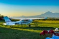 A light single-engine aircraft stands at a small rural airfield covered with green grass against a mountain landscape.