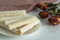 On a light table, sweets for tea and coffee on a porcelain plate. Wafer rolls with coconut flakes and cream next to a bouquet of