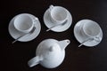 Light tea dishes are placed on a dark table. A white teapot and three mugs with saucers and spoons Royalty Free Stock Photo