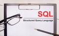 On a light wooden background glasses, a pen and a sheet of paper with the text SQL Structured Query Language. Business concept