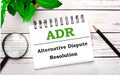 On a light wooden background, a magnifying glass, a pencil, a green plant and a white notebook with text ADR Alternative Dispute Royalty Free Stock Photo