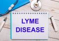On a light wooden background there is a stethoscope, a blue notebook, a white pen and a sheet of paper with the text LYME DISEASE
