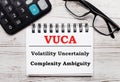 On a light wooden table calculator, glasses and a blank notepad with the text VUCA Volatility Uncertainly Complexity Ambiguity. Royalty Free Stock Photo