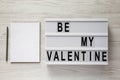 Lightbox with text `Be my Valentine`, blank notepad over white wooden surface. Valentine Day 14 February