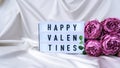 Lightbox with text HAPPY VALENTINES Bouquet of beautiful violet peonies on white silk fabric background. Copy space. Greeting card