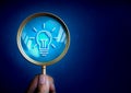 Lightbulb icon in magnifying glass lens in hand on dark blue background with many question mark symbol. Royalty Free Stock Photo