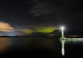 The lighthouse shines at night on the shore of the lake against the background of the starry sky and the aurora borealis