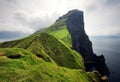 lighthouse sitting on top of a grassy hill close to a rocky black cliff looks over the tranquil sea on cloudy day. Breathtaking Royalty Free Stock Photo