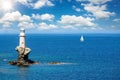 A lighthouse on a steep rock in calm, blue sea with a passing by sailing boat Royalty Free Stock Photo