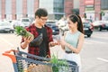 Smiling asian couple standing near shopping cart,man holds pineapple and cheerful woman counts the banknotes