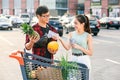 Likable asian couple standing near shopping cart,man holds pineapple and melon and beautiful woman counts the banknotes