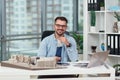 Likable smiling successful bearded 35-aged architect in glasses sits at his workplace with mock-up of future buildings.