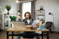 Likable young caucasian man sitting at desk with laptop and holding papers.