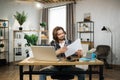 Likable young caucasian man sitting at desk with laptop and holding papers.