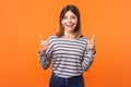 Like, thumbs up! Portrait of joyful kind young woman with brown hair in long sleeve striped shirt. isolated on orange background