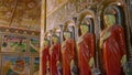 Line of Buddhist statues in ornate temple. Golden walls, intricate murals scenes. Robed figures stand serene, symbols of