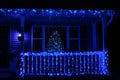 A lit Christmas tree in front of a window framed by a lighted front porch and roofline with blue, purple and green lights