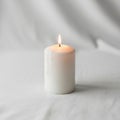 A lit, cylindrical white candle is placed on a soft white fabric surface Royalty Free Stock Photo