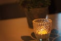 Lit tealight candle in an intricate mosaic-like glass cup or candle holder on the table. Green potted houseplant in the background