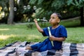 Little adorable African girl in blue dress, blowing and enjoying soap bubbles in park or garden, sitting on checkered Royalty Free Stock Photo
