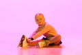 Little adorable baby girl, chide in sweater and skirt, sitting on floor and tying laces on skates against pink