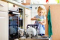 Little adorable cute toddler girl helping to unload dishwasher. Funny happy child standing in the kitchen, holding