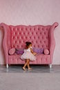 A little African-American girl in a pink dress is standing near the sofa with her back to the camera, unpacking a gift Royalty Free Stock Photo