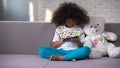 Little African female kid sitting on sofa playing on cellphone, gadget addiction