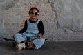 Little African kid boy with dreadlock hairstyle wear cool gold sunglasses, necklace chain, jeans bib and sneaker smiling