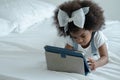 Little African kid girl with bow tie on afro hair is lying on white bed and using tablet computer for online learning at home Royalty Free Stock Photo
