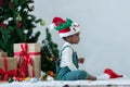 Little African kid girl smile in Santa hat and green clothes sitting with gift boxes, toys and Christmas tree with decorated balls Royalty Free Stock Photo