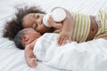 Little African older sister sucking milk from bottle, hugging and take good care of her newborn baby brother lying on bed at home Royalty Free Stock Photo