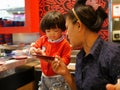 Little Asian baby girl enjoys picking raw ingredients, for her mother, for hotpot cooking, serving on a moving conveyor belt Royalty Free Stock Photo