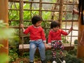 Little Asian baby girls, sisters, enjoy being in a swing together in a garden, with their mother watching close by