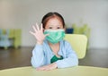 Little Asian child girl wearing a protective mask with showing five fingers sitting on kid chair in children room Royalty Free Stock Photo