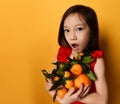 Asian child in red blouse. Looking wondered, holding an armful of tangerines and oranges, posing on orange background. Close up