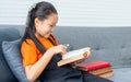 Little Asian cute girl wearing casual clothes, sitting on sofa in indoor comfortable living room at home in weekend, reading book Royalty Free Stock Photo