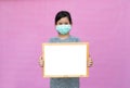 Little asian girl in a protective medical mask holding whiteboard isolated on pink background.