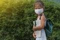 Little asian girl wearing face mask with green back pack holding stationery in green nature park background. Back to school Royalty Free Stock Photo