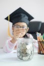 Little Asian girl wearing graduate hat putting the coin into clear glass jar piggy bank and smile with happiness for money saving Royalty Free Stock Photo