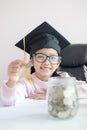 Little Asian girl wearing graduate hat putting the coin into clear glass jar piggy bank and smile with happiness for money saving Royalty Free Stock Photo